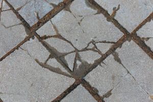 A close-up of gray concrete pavers with dark, cracked grout lines and surface fissures.