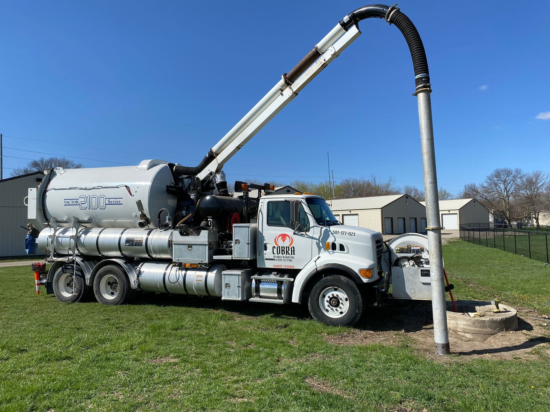 White vacuum truck pumping material through a long hose into an upright pipe on a grassy site