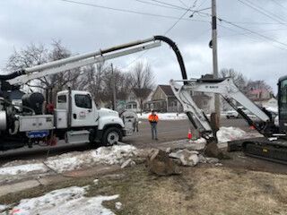 Utility trucks and workers repairing a road with a large black pipe on a muddy street.