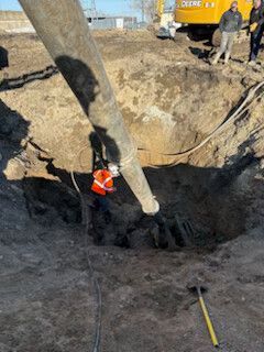Worker in orange safety vest standing in a deep trench beside a large pipe and excavator.