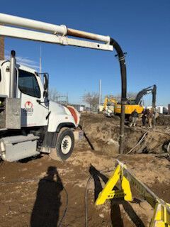 Construction truck with white boom over a trench, yellow excavator in the background on a dirt site.