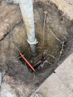 Open excavation with a vertical pipe and red tool or rod at the bottom of a dirt hole.