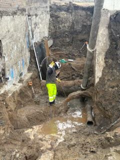 Worker in neon pants stands in a muddy excavation trench with exposed pipes and soil walls.