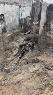 Muddy construction trench with exposed pipes and a central wooden support beam against concrete walls