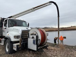 White utility truck pumping water through a hose near a river, with two workers in orange safety gear nearby