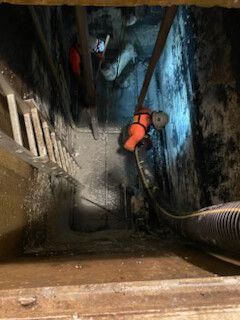 Worker in orange safety gear descending a narrow, steep tunnel beside a large pipe.