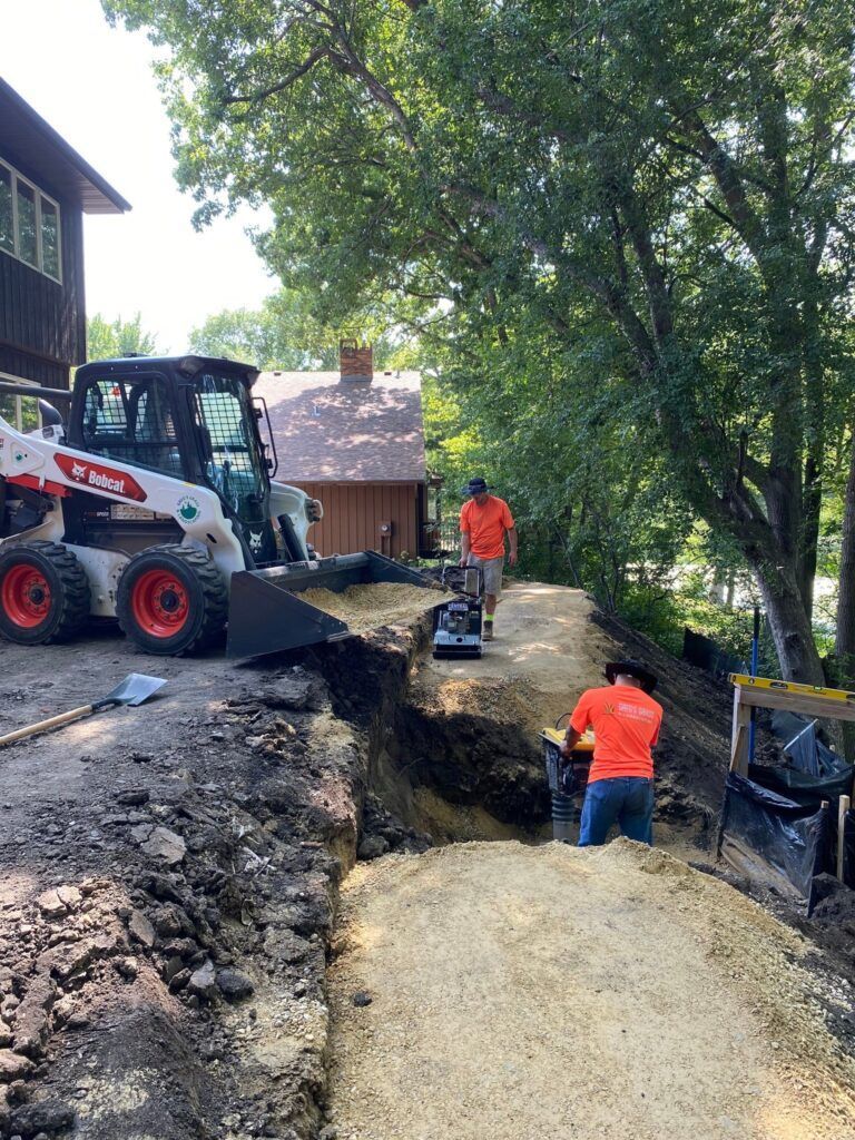 Two workers in orange shirts work on a gravel path near a house, using a Bobcat skid steer and a compactor.