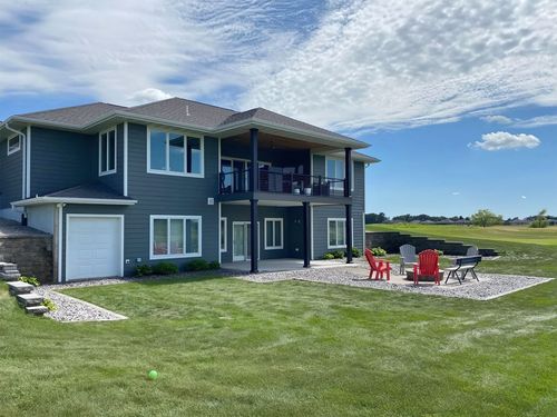 A two-story blue house with a gravel patio, red chairs, and a fire pit, overlooking a large green lawn under a blue sky.