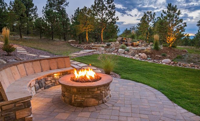 Circular brick patio with fire pit, back wall, and two pillars in backyard setting.