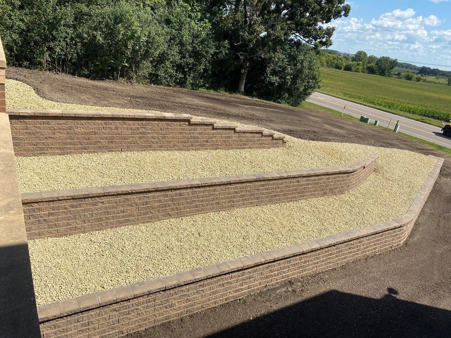 A landscape with terraced retaining walls built from tan stacked stones, filled with light-colored gravel, beside a road.