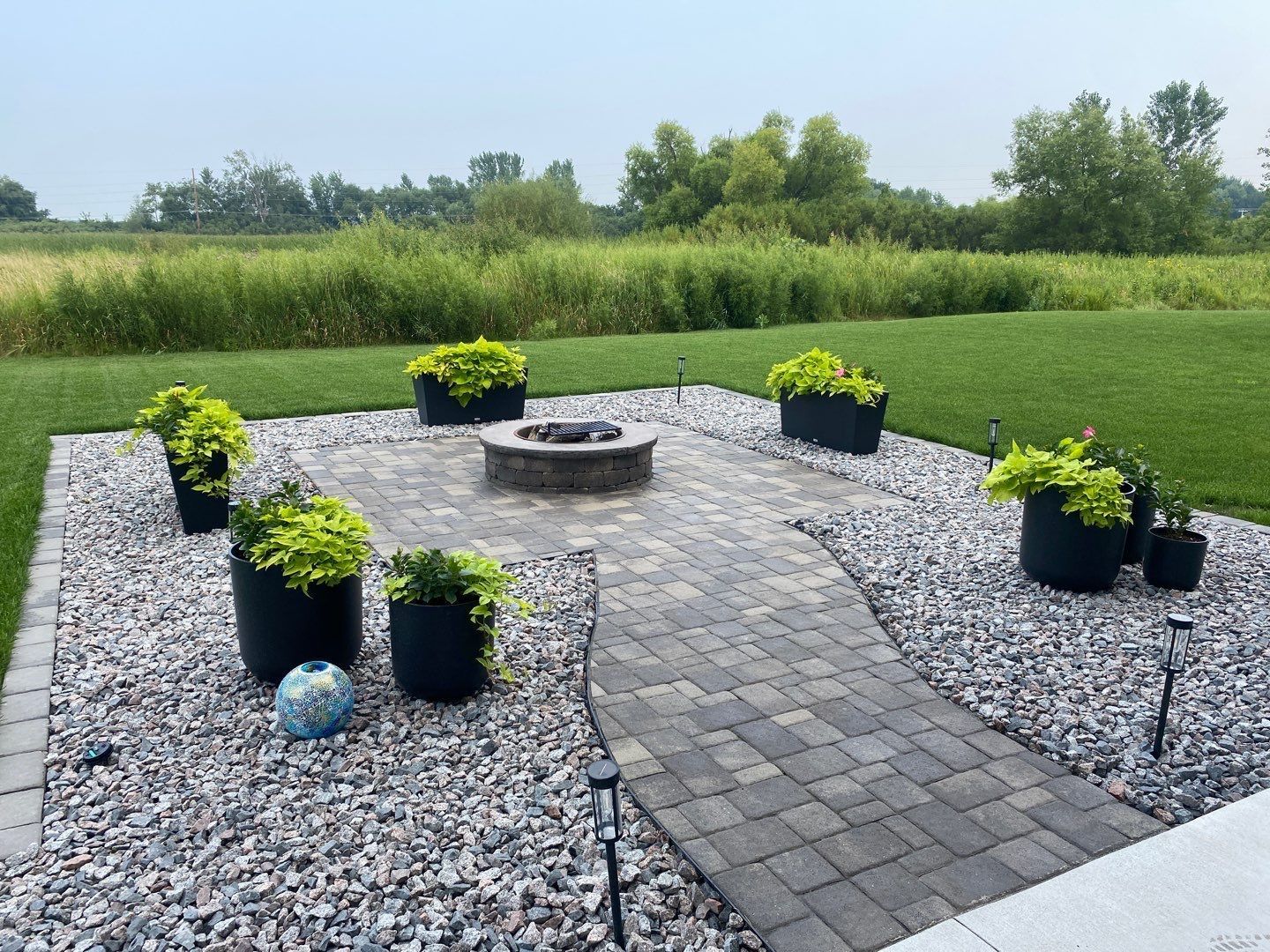 A circular stone fire pit sits in a paved patio surrounded by gray gravel, potted green plants, and a grassy backyard.