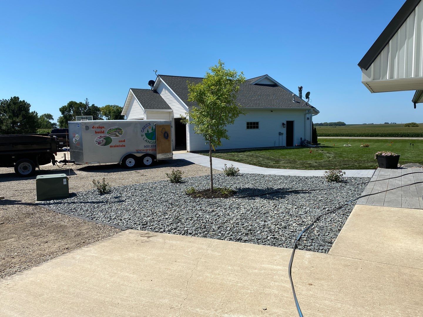 A white building with a gray roof and a parked trailer sit behind a landscaped garden bed with a small tree on a sunny day.
