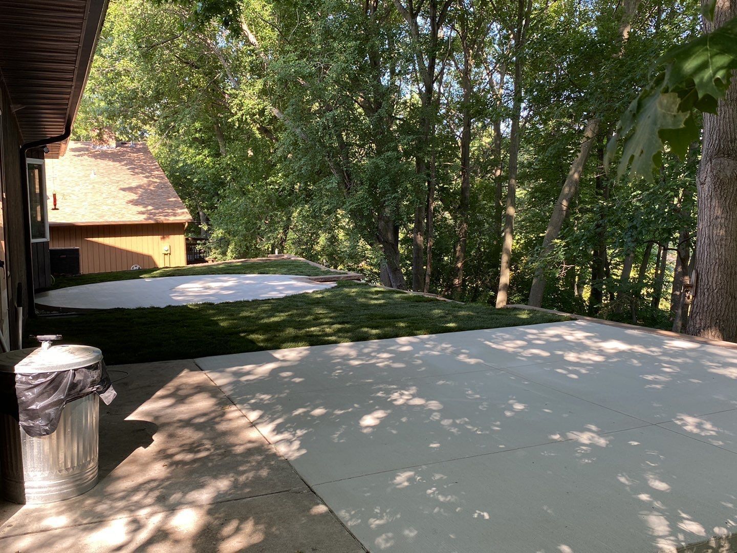 A concrete patio and grassy area sit beside a house, leading to a tree-lined wooded slope in the background.