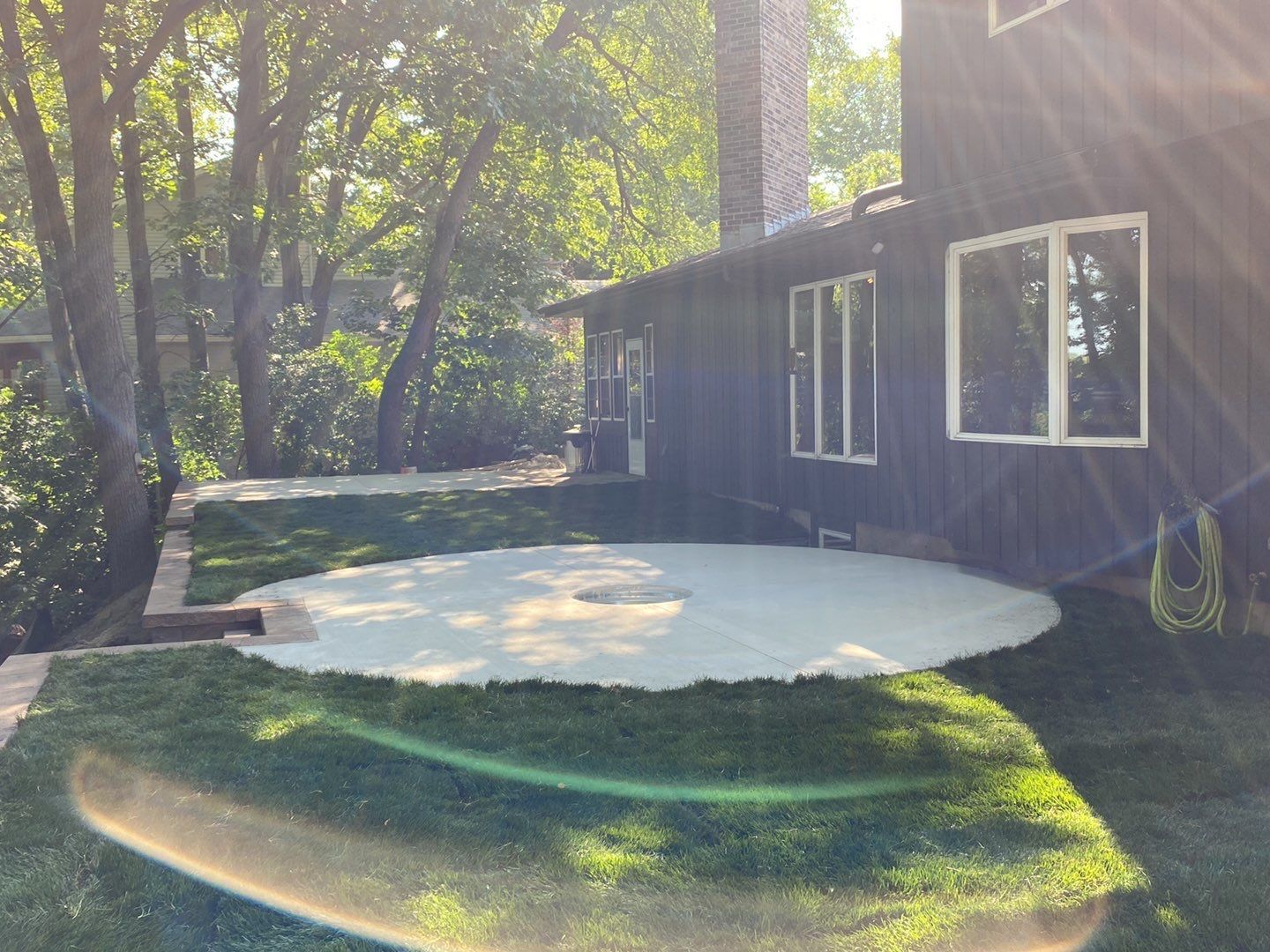 A circular concrete patio sits in a backyard next to a dark-sided house, surrounded by lush green lawn and shade trees.