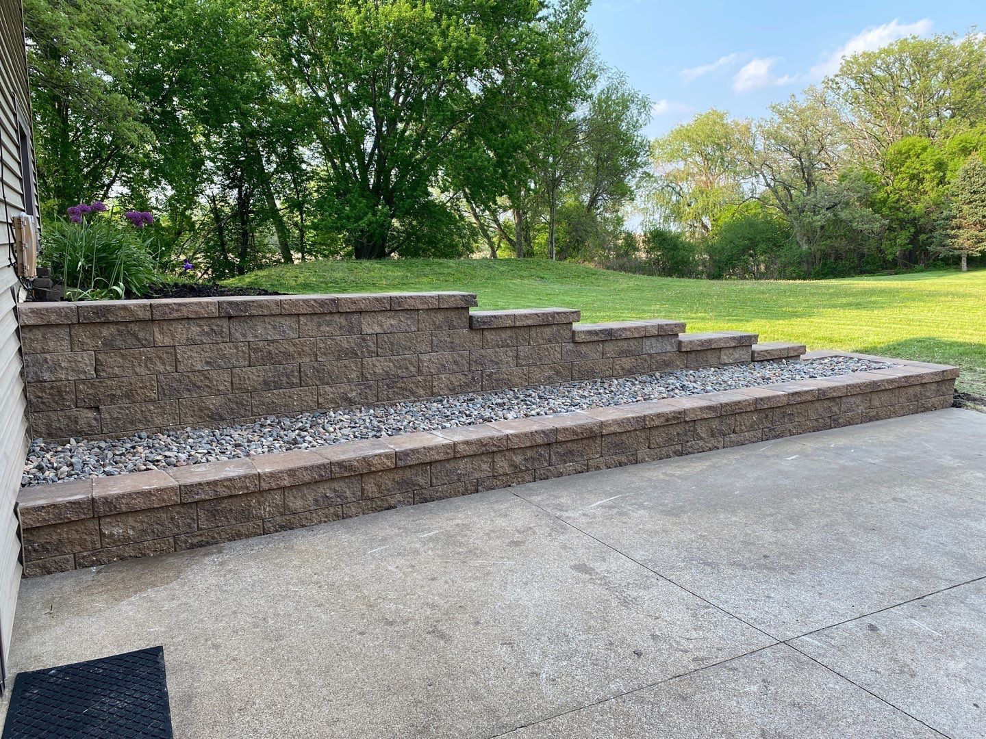 A tiered, brown stone retaining wall borders a concrete patio, with gravel in front and green trees in the background.