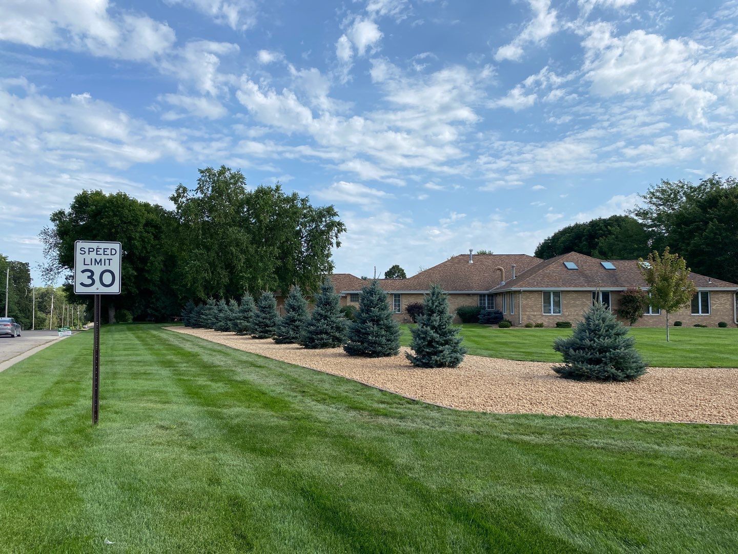 A 30 mph speed limit sign stands beside a green lawn and a row of evergreen trees in front of a brick house.