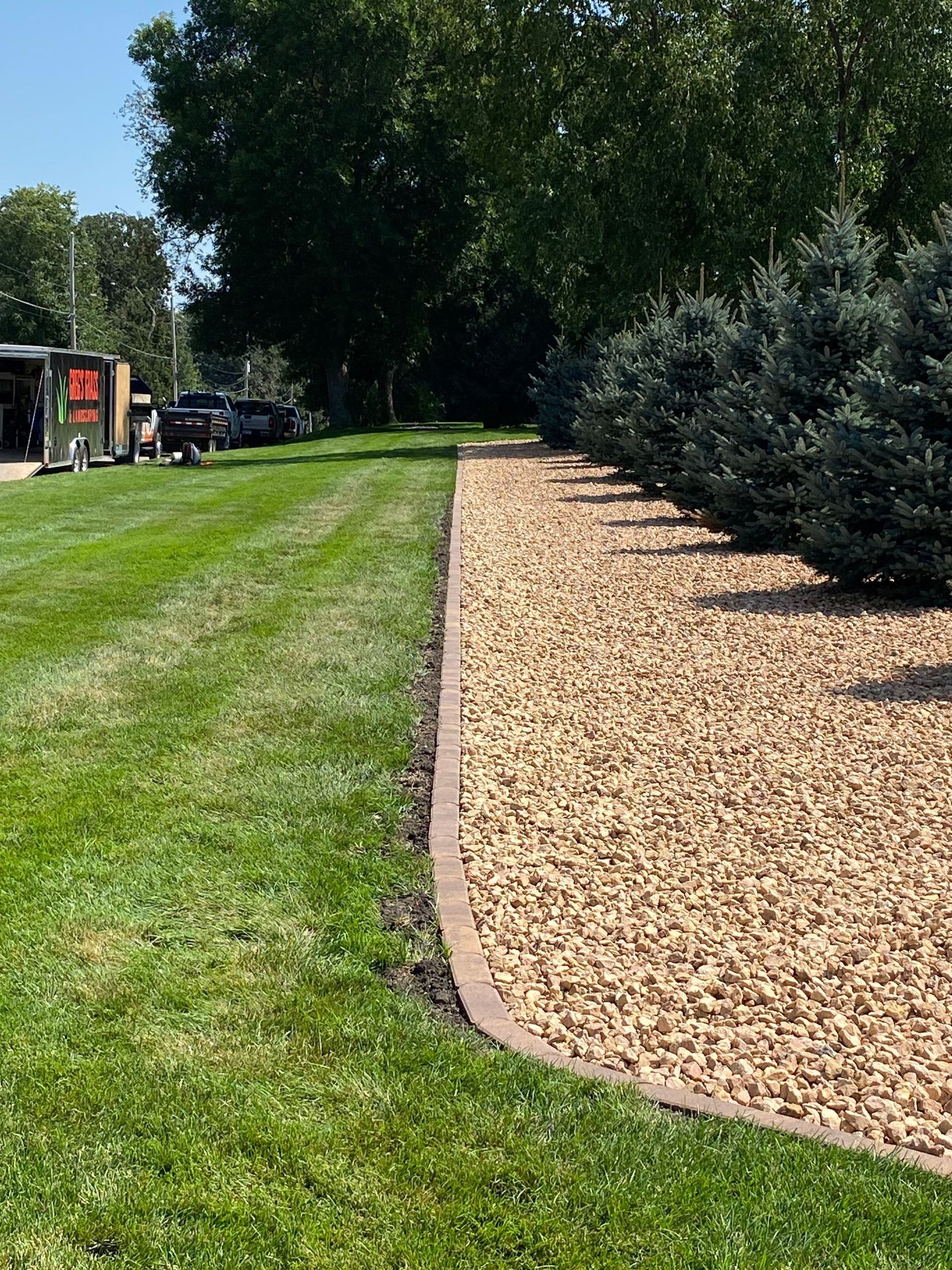 A landscaped garden bed with tan gravel and a curved concrete border, sitting next to a manicured green lawn and pine trees.