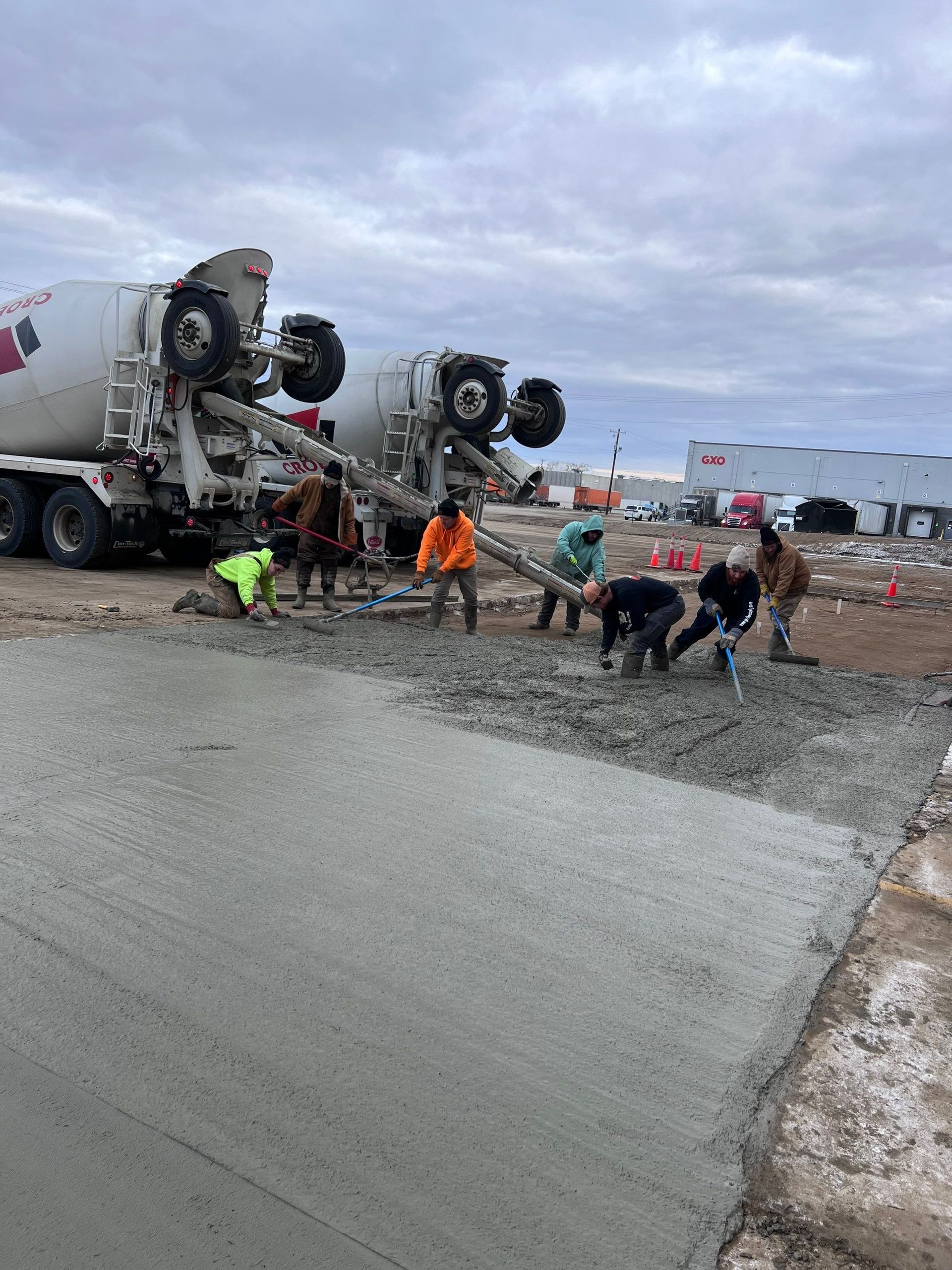 Construction workers spreading wet concrete from mixer trucks onto a job site under a cloudy sky.