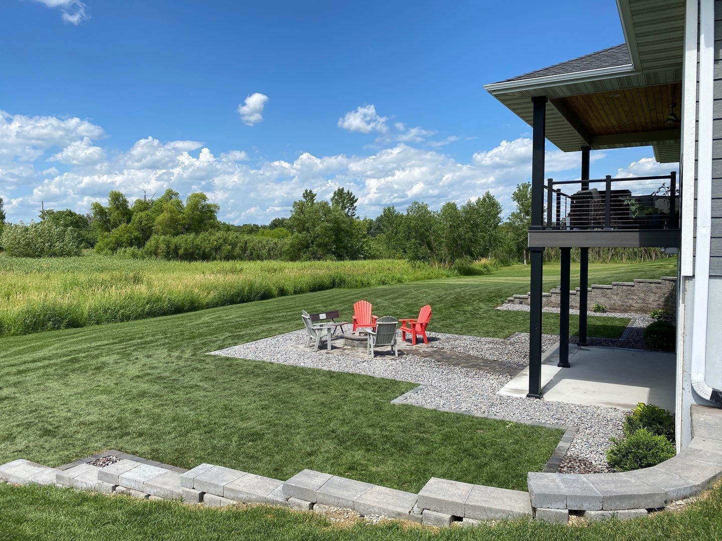 A gravel patio with three red Adirondack chairs sits in a backyard next to a house with a deck overlooking a field.