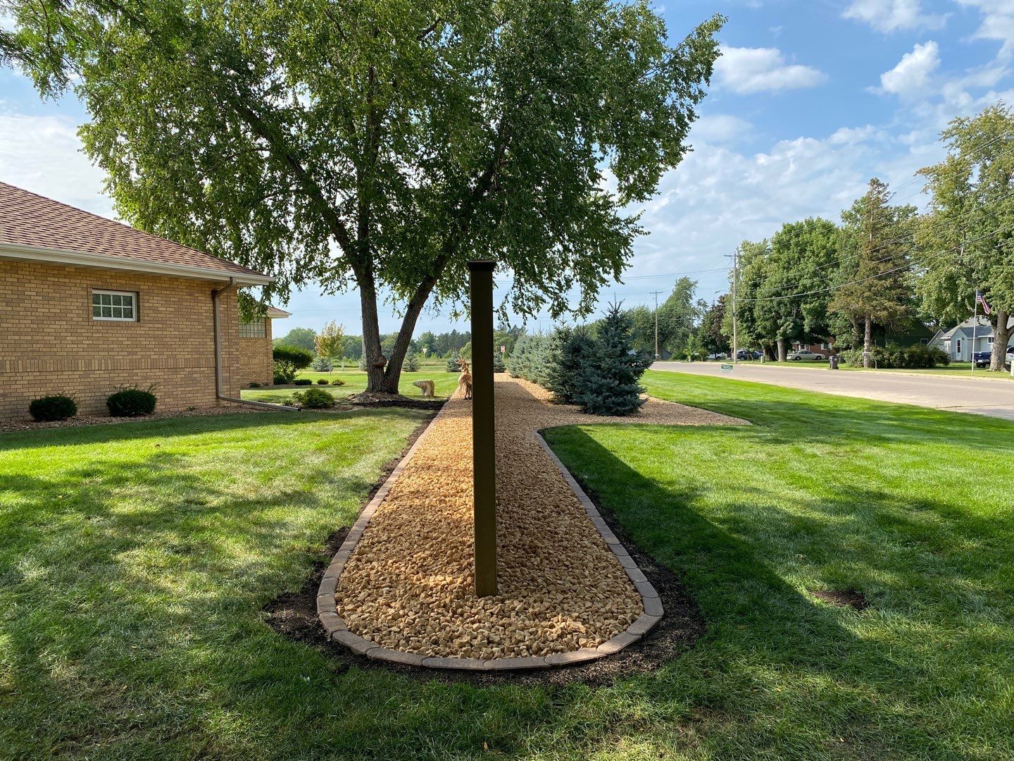A gravel flower bed with a central wooden post sits in a green lawn, adjacent to a brick building and leafy trees.
