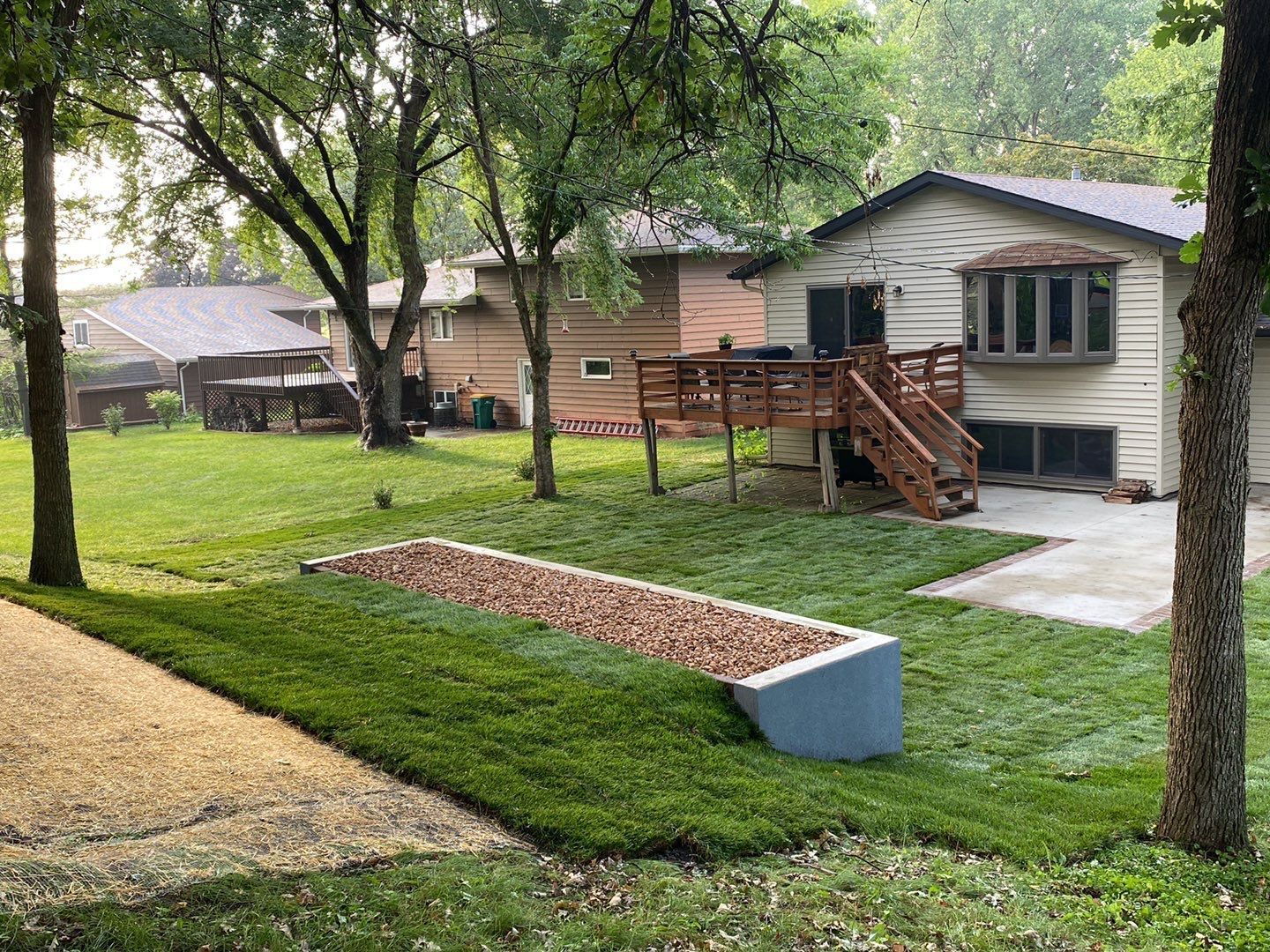 A backyard features a freshly sodded lawn, a tan house with a wooden deck, and a raised concrete planter filled with mulch.