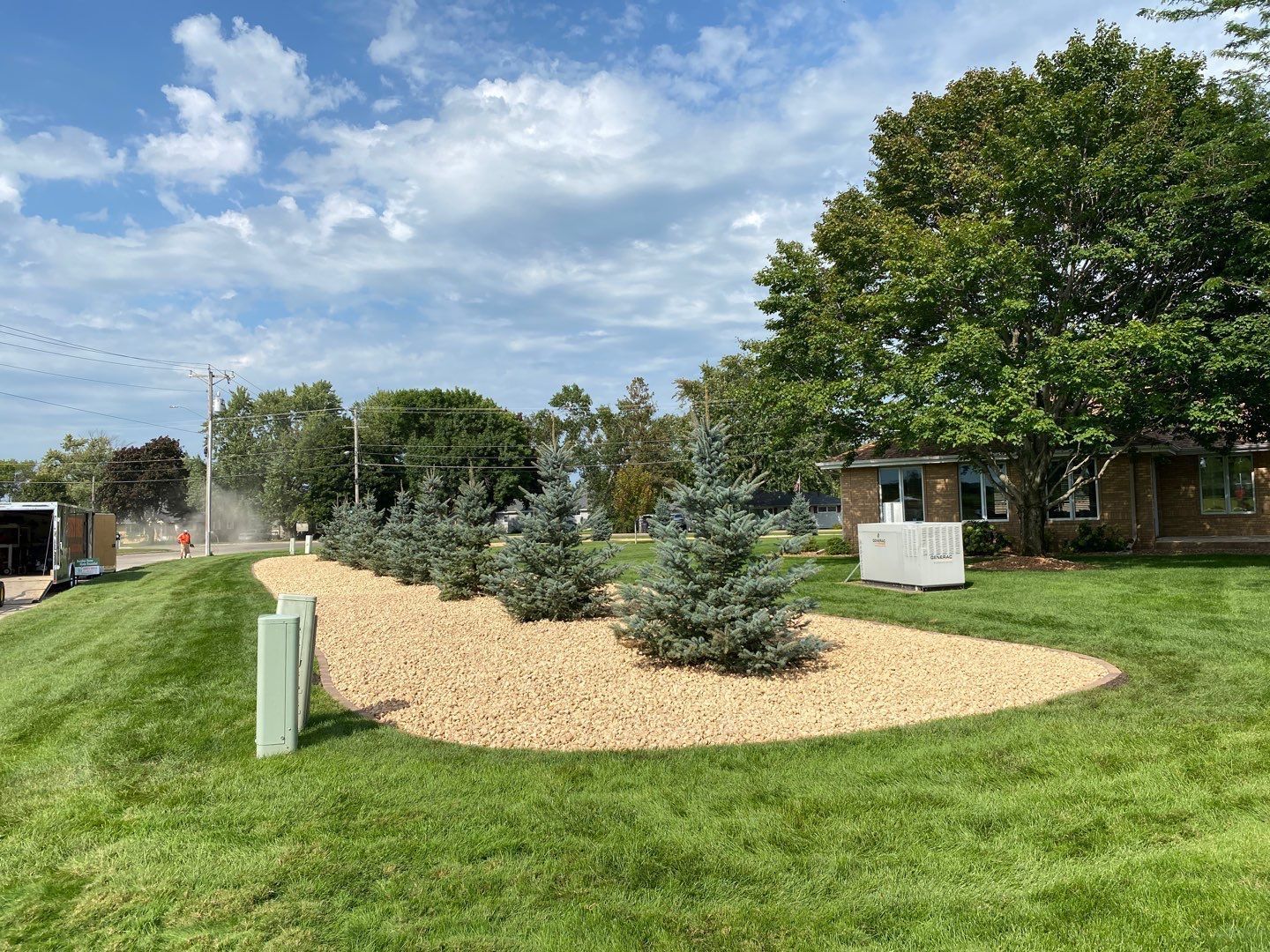 A gravel landscape bed features a row of blue spruce trees, surrounded by mown green grass under a partly cloudy sky.