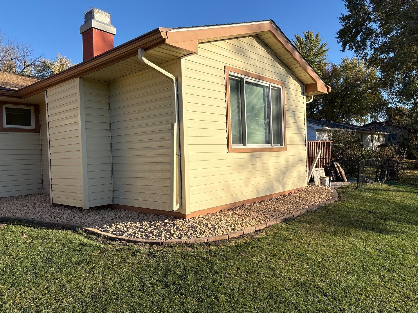 A light yellow house exterior with a brick chimney, a single window, and a gravel garden border under a clear blue sky.