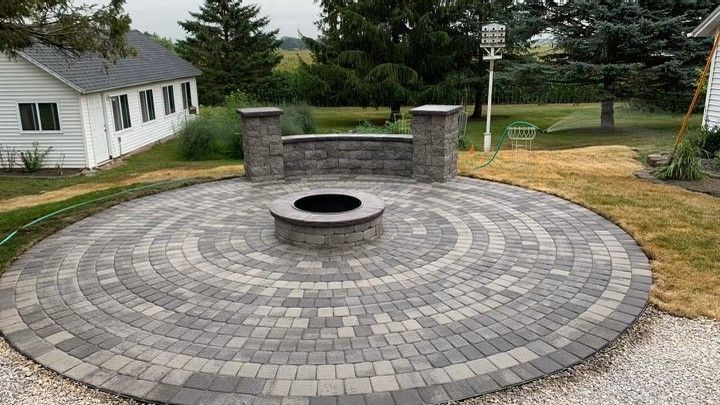 A circular gray paver patio featuring a central stone fire pit and a curved seating wall with two stone pillars.