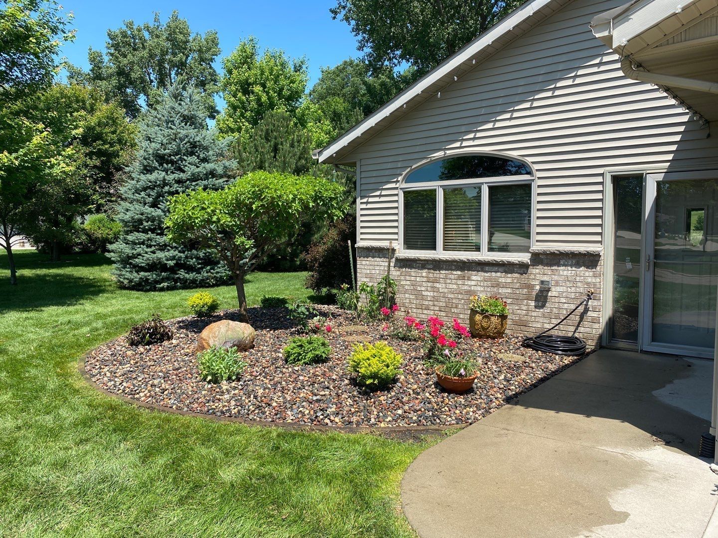 A stone-mulched garden bed with small shrubs and pink flowers sits next to a light-sided house under a clear blue sky.