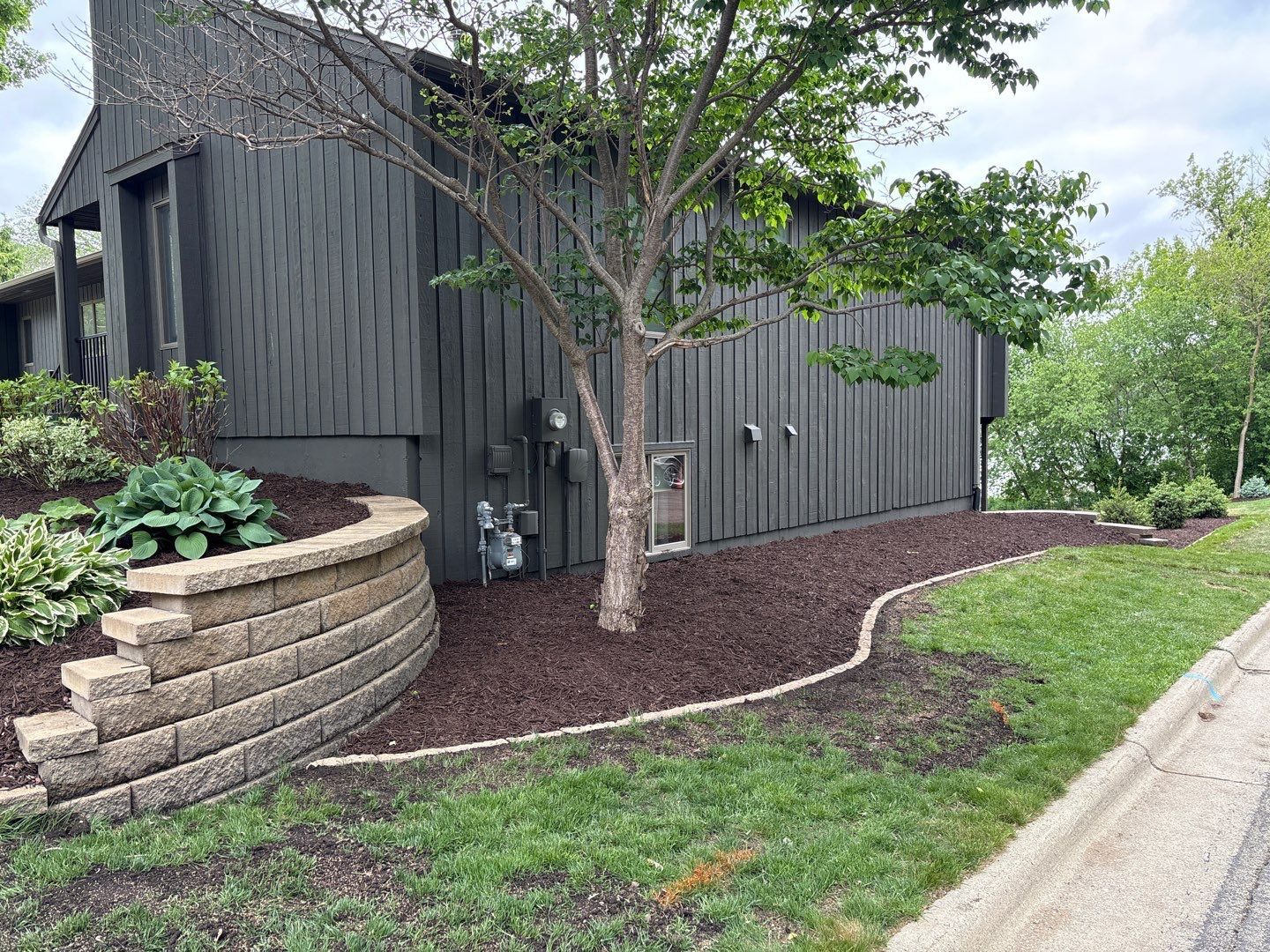 Dark gray house with landscaped front yard, tree, stone retaining wall, and driveway edge