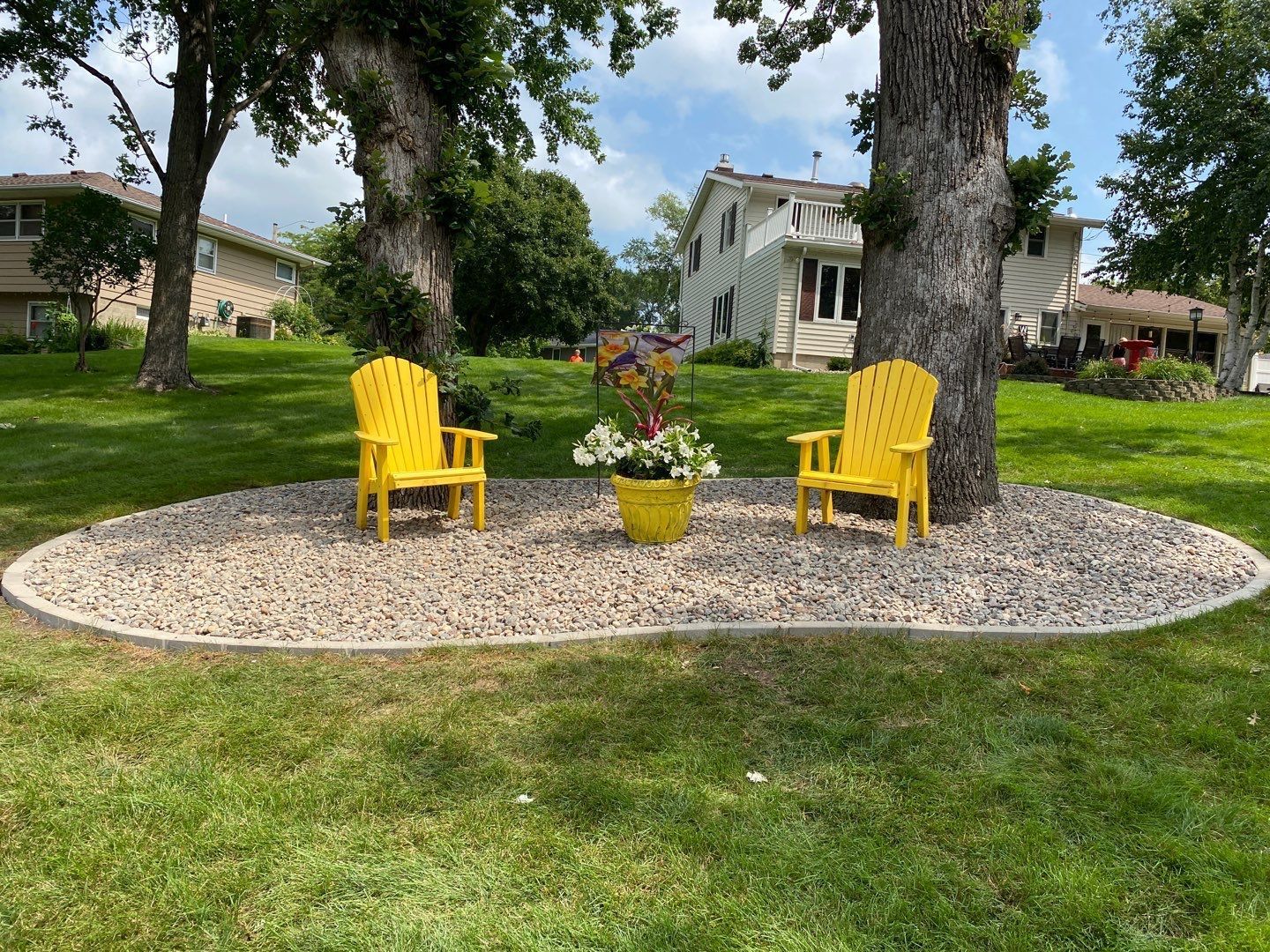 Two yellow chairs around a flower pot on a pebble patio in a green yard