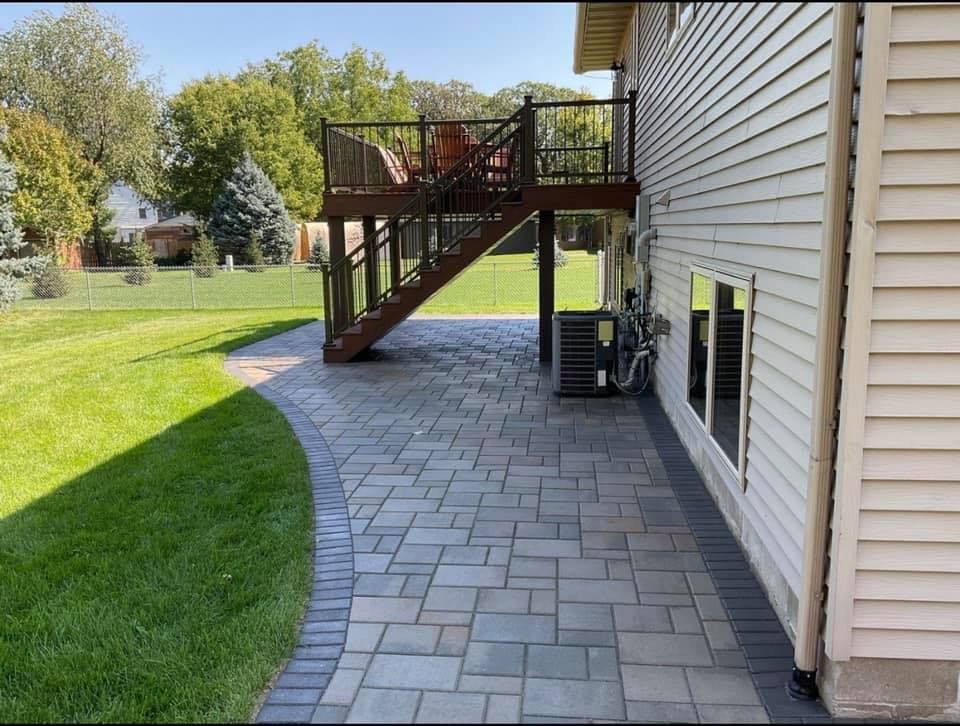 Paved patio beside a house with wooden deck stairs and green lawn.