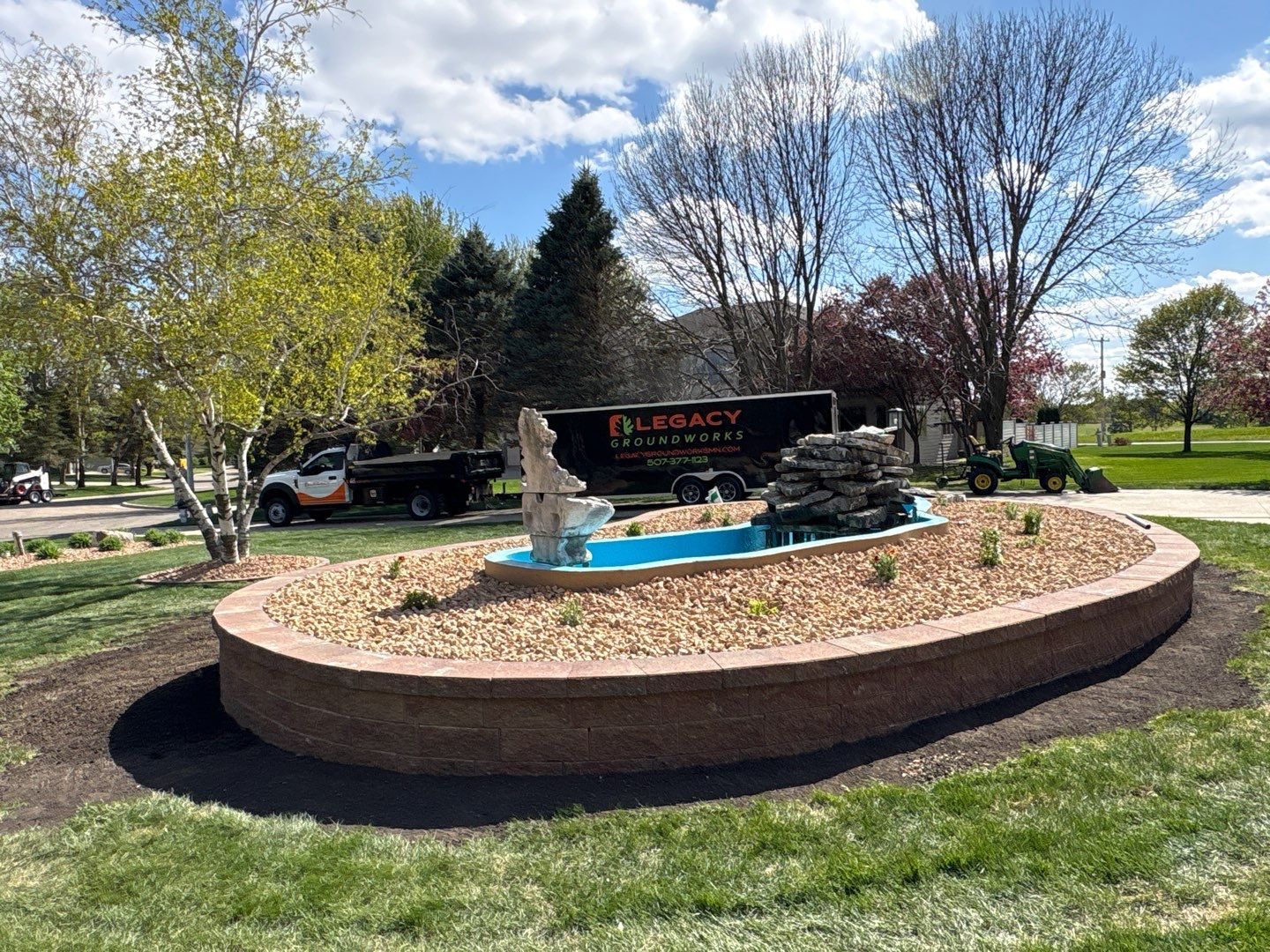 Round brick fountain with turquoise water in a landscaped park, with trees and a sign in the background