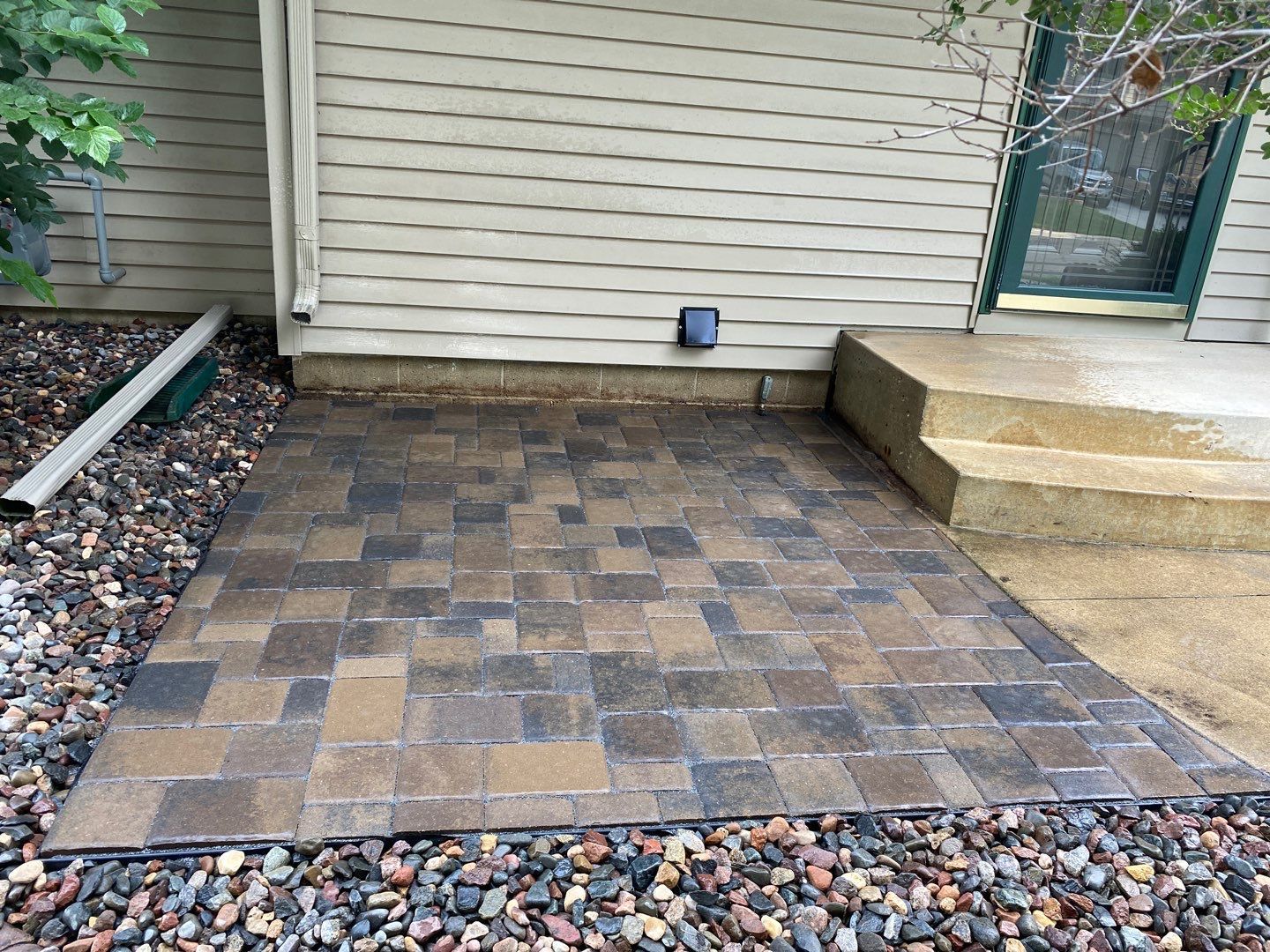 Small paver patio beside house wall, bordered by stone and gravel, next to concrete steps and a door