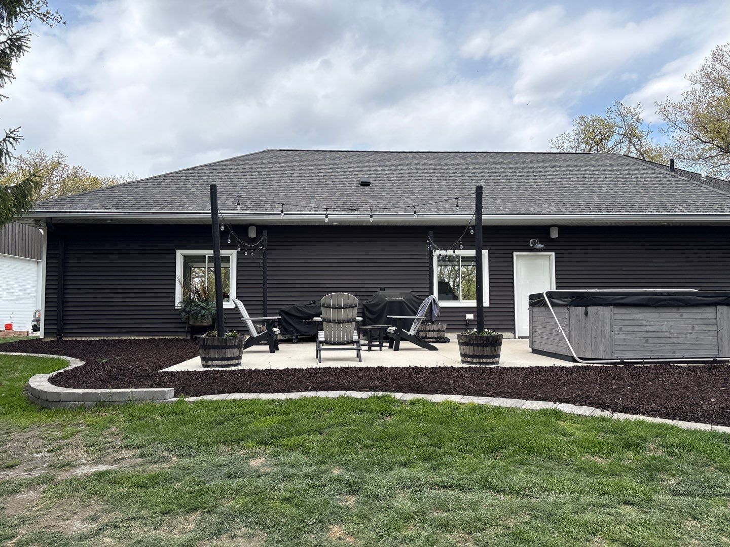 Black house with patio chairs, hot tub, and curved stone path under a cloudy sky.