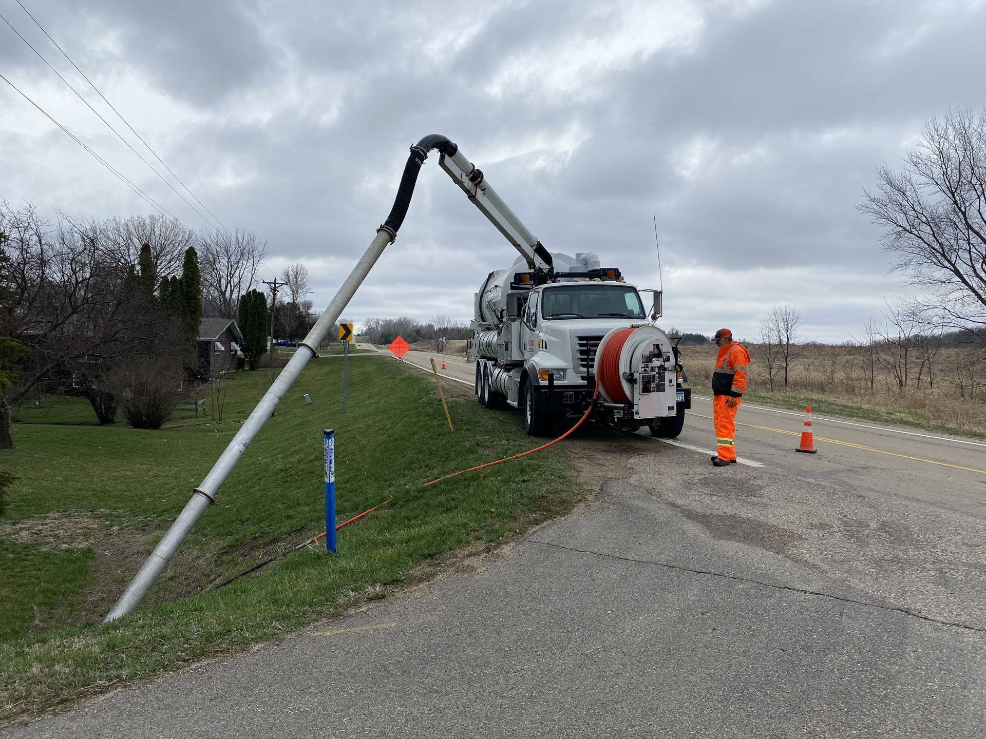 Utility truck with boom arm on rural roadside, workers in orange safety gear, and traffic cones marking the area