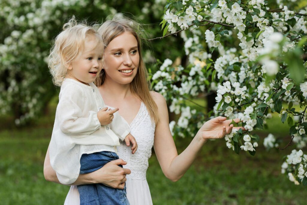 A person in a white dress holds a toddler in a blooming orchard, gently touching the white flowers.