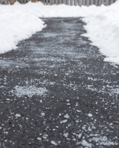 A paved path covered in scattered ice and snow, with thick mounds of snow lining both sides of the walkway.