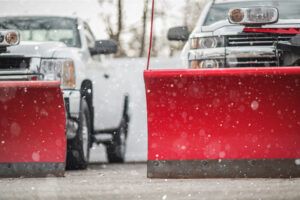 Two white pickup trucks with large, bright red snowplows attached to the front during a snowfall.