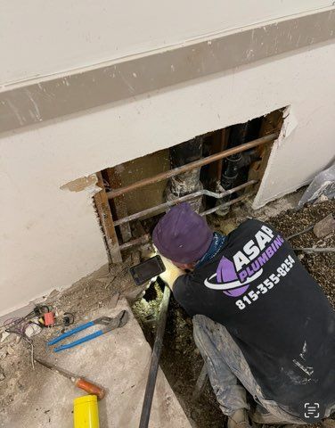 Plumber working on pipes in wall, wearing a purple hat and black shirt.