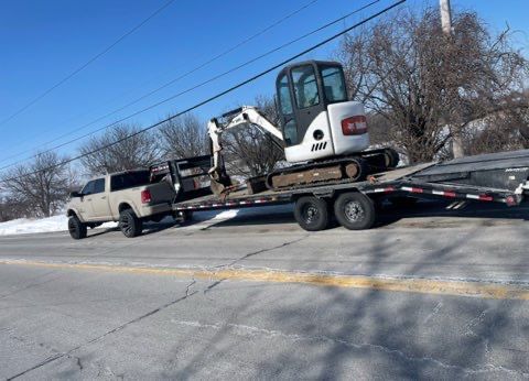 A light-colored pickup truck towing a flatbed trailer with a Bobcat excavator on a road.