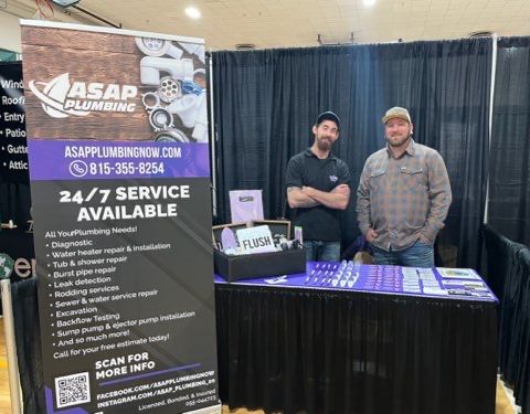 Two men at a plumbing services booth at an event. Signage advertises 24/7 service.