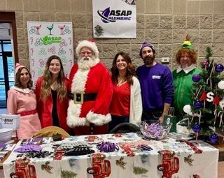 People dressed in festive attire at a holiday event. Santa stands with others at a decorated table.