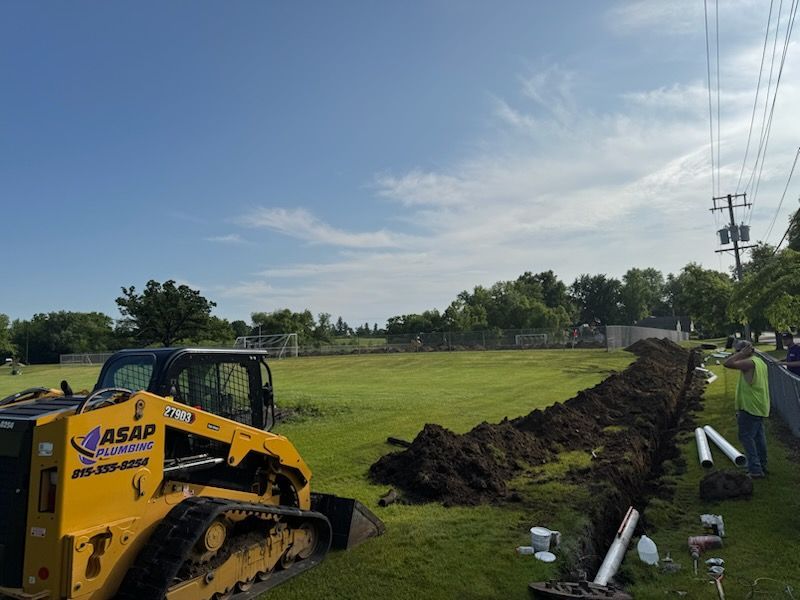 A yellow track skid steer digs a trench in a grassy field near a road. White pipes are nearby.