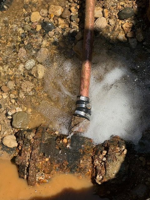 Water spraying from a copper pipe joint connected to a rusty pipe in a dirt and gravel area.