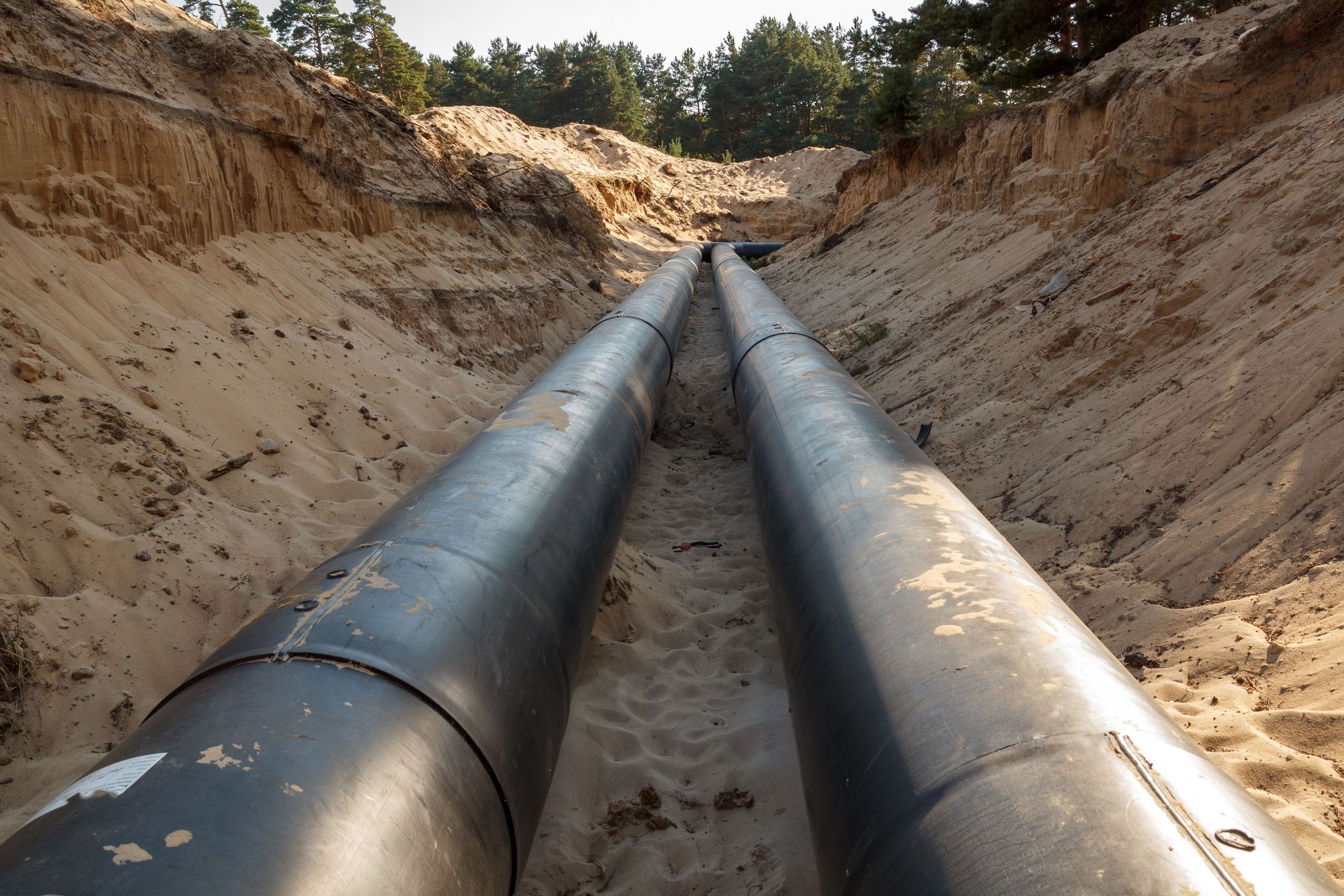 Two large black pipes running side-by-side in a sand-filled trench, under a forest.