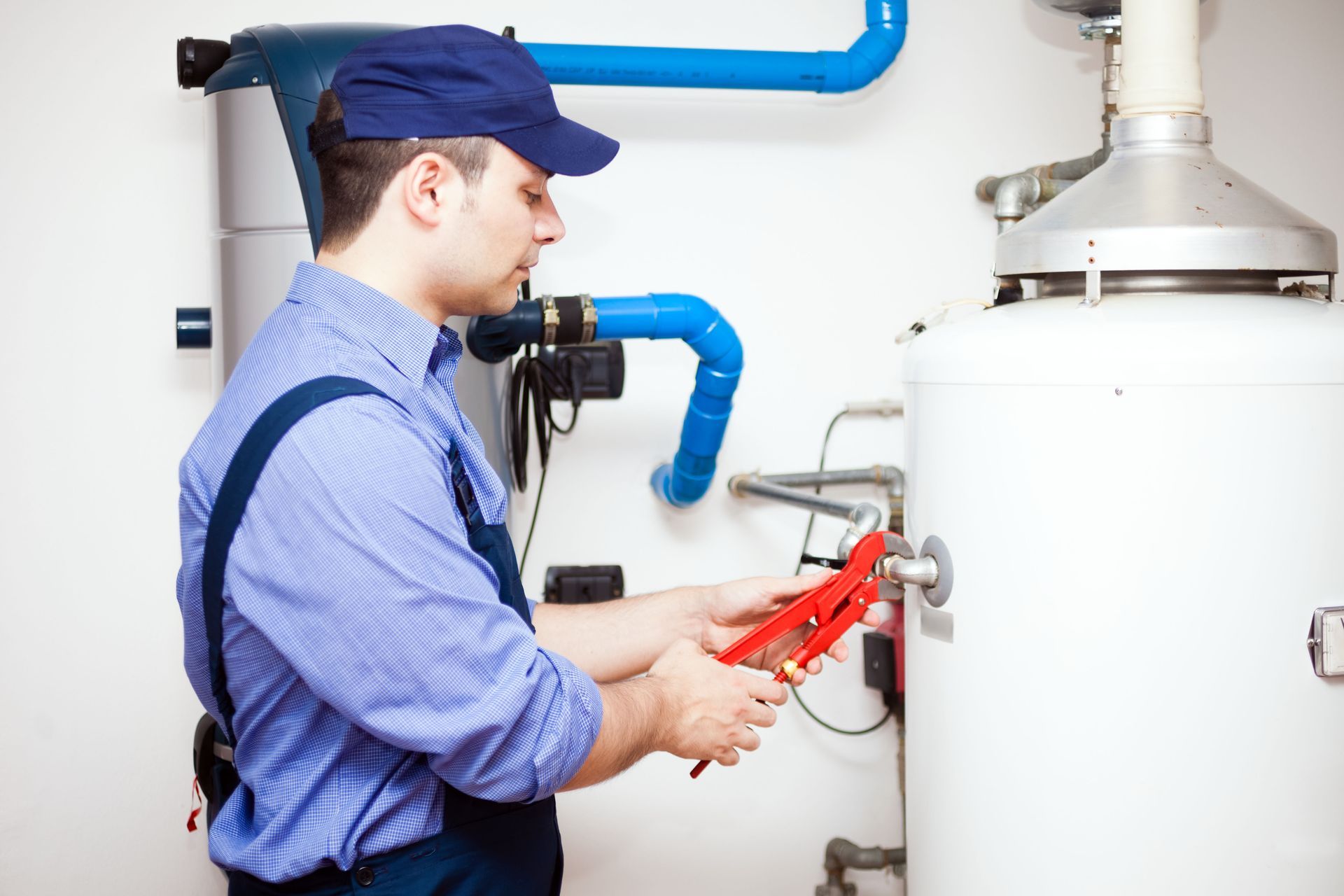 Plumber in blue shirt and cap using pliers to work on a white water heater.