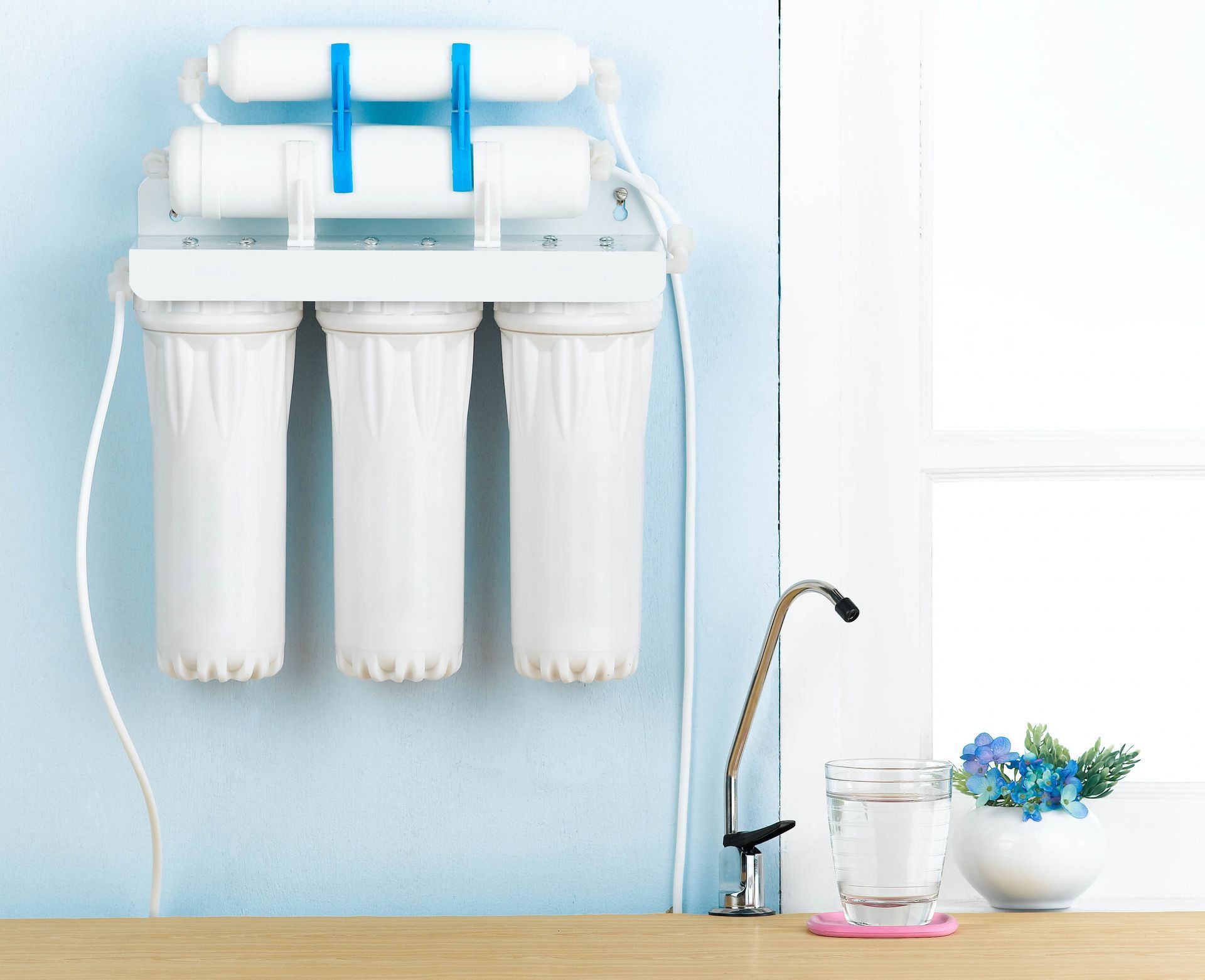 White water filter system mounted on a light blue wall with a faucet, glass of water, and flowers.