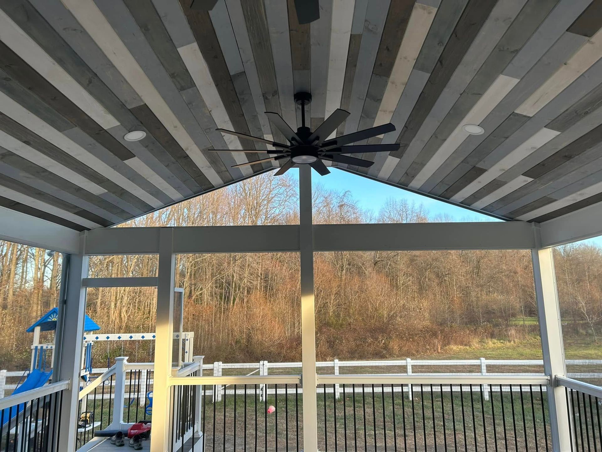Covered porch with wood-plank ceiling, ceiling fan, and view of playground and trees.