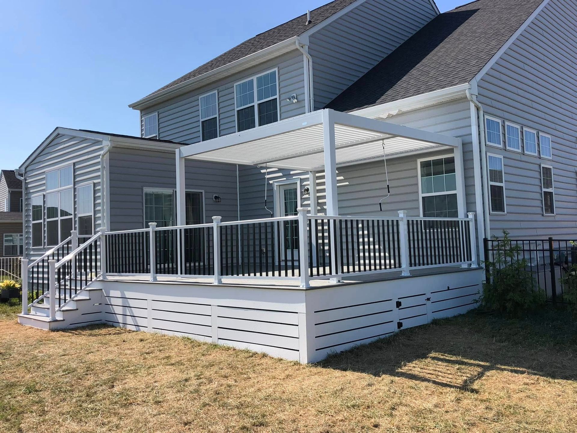 Back of a house with a white deck, black railing, and pergola under a blue sky.